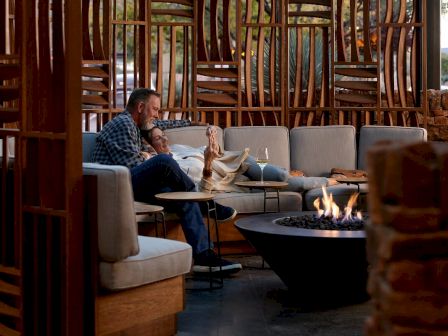 A couple is relaxing on a couch in a cozy, outdoor area near a fire pit, surrounded by modern wooden and stone decor, with drinks in hand.