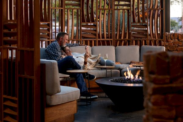 A couple is relaxing on a couch in a cozy, outdoor area near a fire pit, surrounded by modern wooden and stone decor, with drinks in hand.