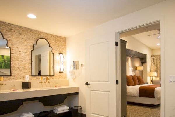 The image shows a hotel room with a bathroom featuring double sinks and ornate mirrors, leading to a bedroom with a large bed and warm lighting.