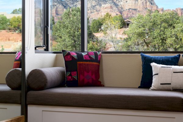 A cozy nook with cushions by a large window overlooks a scenic mountain view, featuring a colorful rug on the wooden floor.