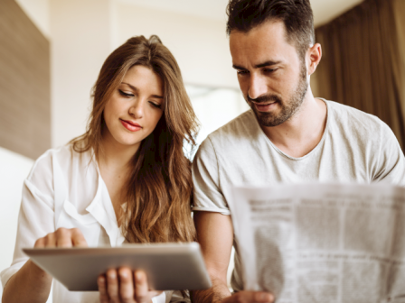 A woman is using a tablet while a man next to her reads a newspaper. They are seated indoors, appearing engaged with their respective devices.