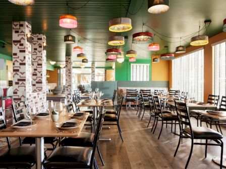 A vibrant restaurant interior with colorful hanging lights, modern decor, neatly arranged tables and chairs, and large windows allowing natural light.