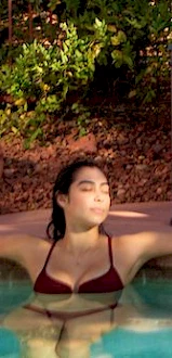 A person is relaxing in a pool, leaning back with arms outstretched, against a backdrop of greenery and rocks.