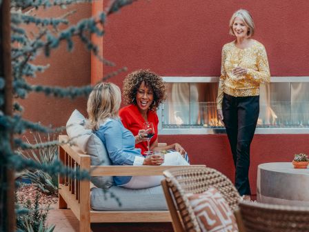 Three women enjoying a conversation and drinks in a cozy outdoor setting with a fireplace and modern furniture.