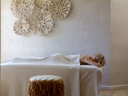 A person is lying on a massage table covered with a white sheet in a room with decorative wall art and a stool. The walls are a neutral color.