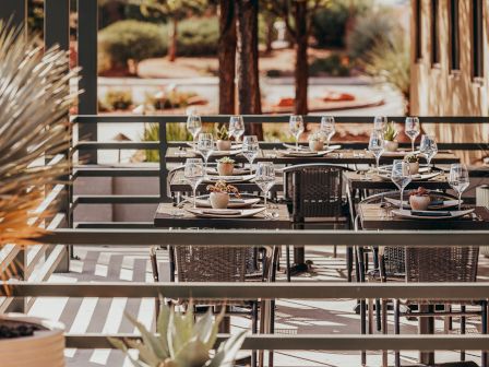 Outdoor dining setup with tables, chairs, and wine glasses on a patio surrounded by greenery under the shade of a pergola.