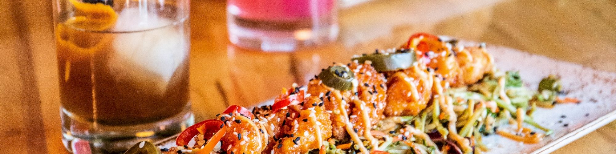 A dish with skewered food on noodles, garnished with peppers and herbs, flanked by pink and brown cocktails. Tacos are in the background.