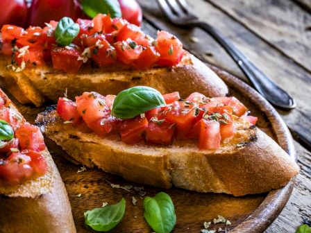 Slices of toasted bread topped with diced tomatoes, basil leaves, and seasoning on a wooden plate with a fork in the background.