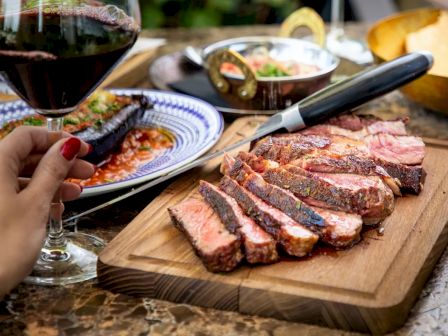 A person holds a glass of red wine next to a cutting board with sliced cooked meat, a knife, and various plated dishes on a table.