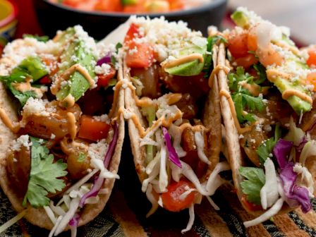 The image shows three tacos filled with various ingredients including diced tomatoes, avocado, cilantro, shredded cabbage, and cheese, placed on a woven mat.