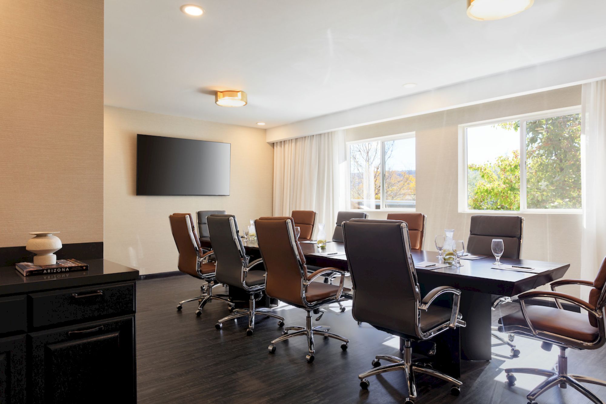 A modern conference room with a long table, eight chairs, a flat-screen TV on the wall, windows, notebooks, and glasses on the table, always ending the sentence.