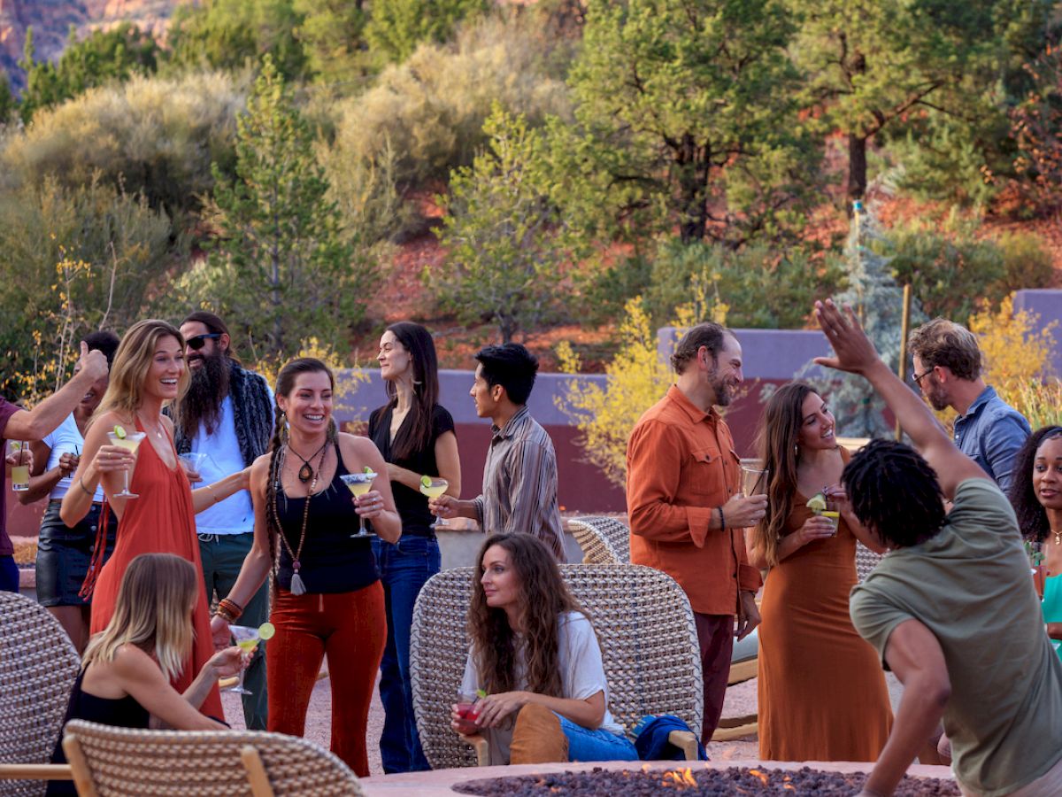 People are socializing at an outdoor gathering in a natural setting, enjoying drinks, conversation, and the scenery, with trees and a rocky backdrop.