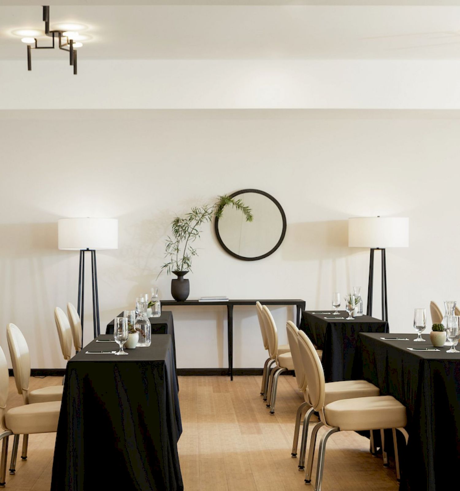 A minimalistic meeting room with tables draped in black cloth, beige chairs, two lamps, a circular wall mirror, and potted plants on a console table.