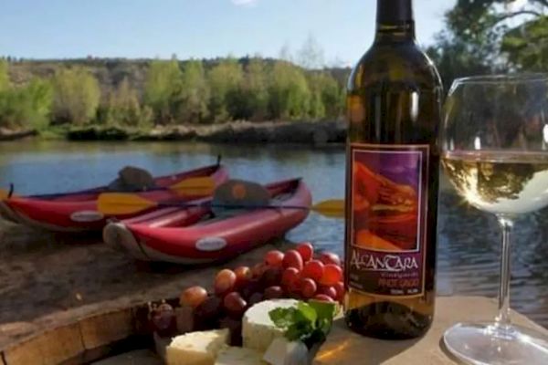 The image shows wine with cheese and grapes on a table near a lake with kayaks.