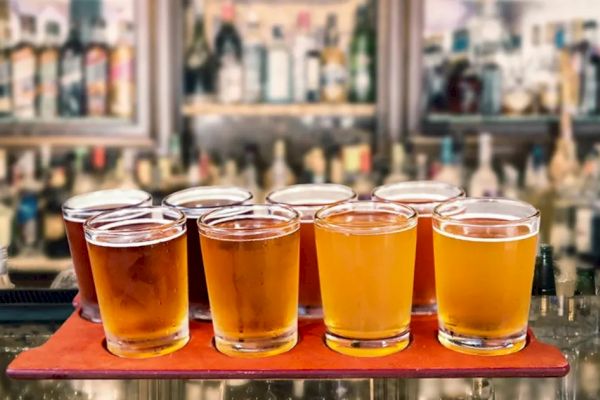 The image shows an assortment of beer glasses filled with different types of beer, placed on a red tray in front of a bar with various bottles.