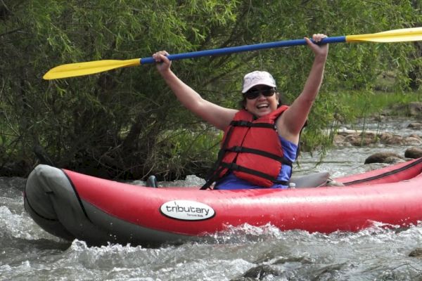 A person in a life jacket and hat is kayaking in a red and gray inflatable kayak on a river, raising their paddle above their head and smiling.