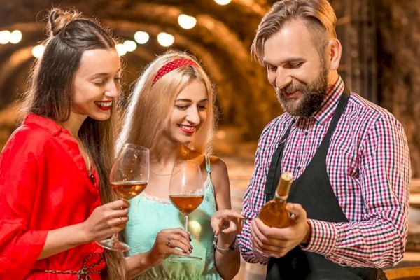 People enjoying a wine tasting in a cellar, holding wine glasses, while a man shows them a bottle of wine, smiling and engaging in conversation.