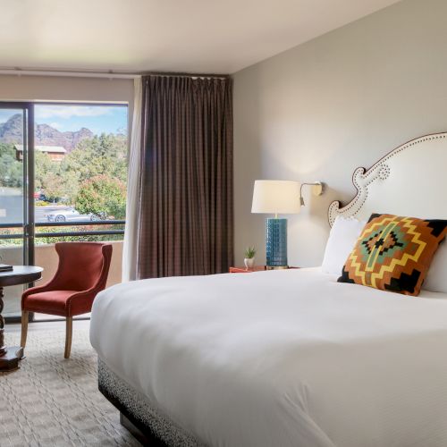 The image shows a hotel room with a large bed, a decorative pillow, two red chairs, a table, and a window offering a scenic view of mountains.