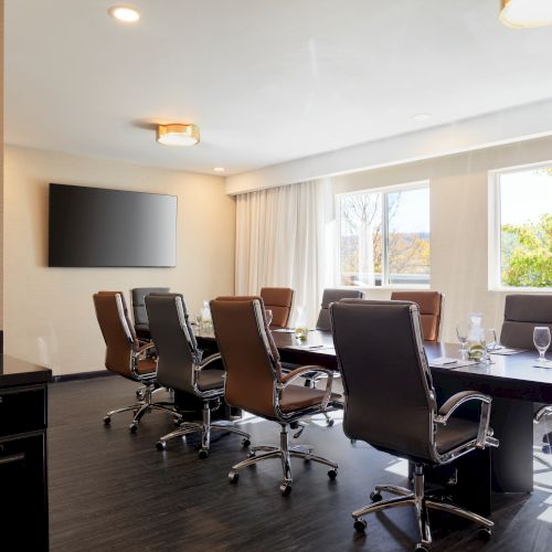 A conference room with a long table, several chairs, a large TV on the wall, and windows letting in natural light.