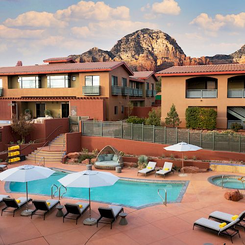 An outdoor pool area with lounge chairs, umbrellas, and a hot tub, surrounded by buildings with a scenic mountain backdrop under a clear sky.