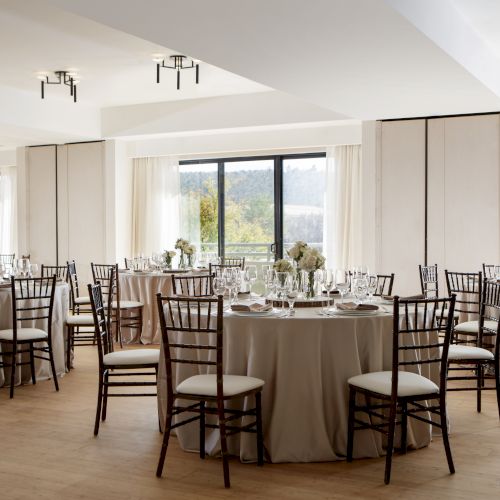 A banquet or event hall with round tables covered in beige tablecloths, black chairs, and white floral centerpieces in a bright room with large windows.