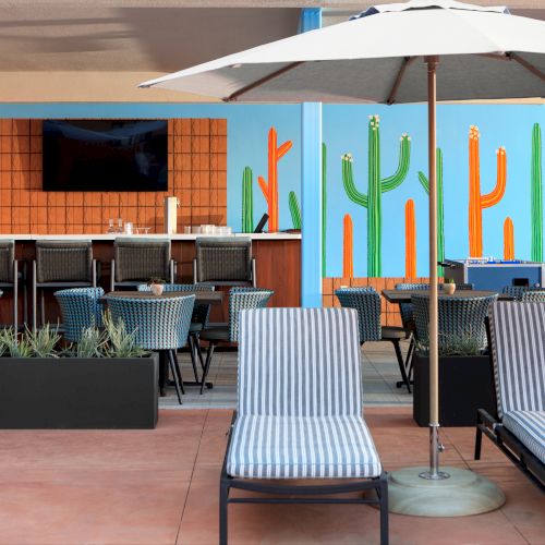 Outdoor dining area with striped chairs, tables, a bar, a large white umbrella, and colorful cactus wall art in the background.