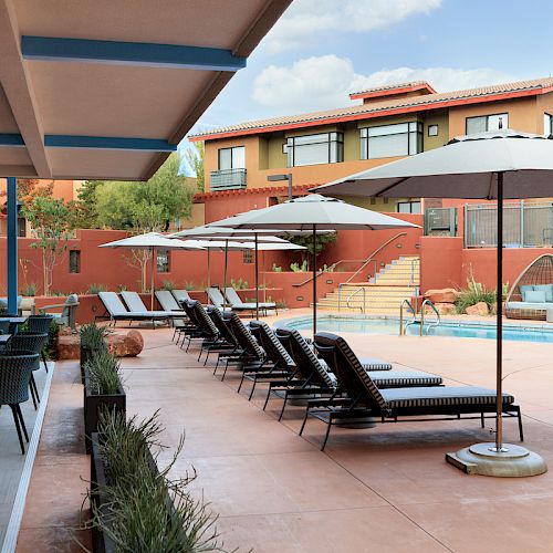 A serene poolside area with lounge chairs, umbrellas, tables, and a building in the background under a partly cloudy sky.