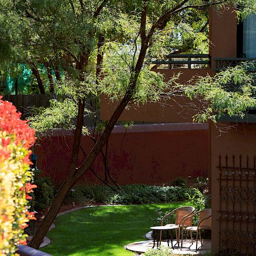 A peaceful garden with green grass, a tall tree, a small table with two chairs, and red flowers. There is a brown building in the background.