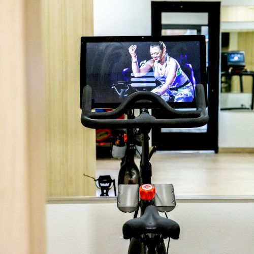 A stationary bike with a screen displaying a fitness instructor is set up in front of a mirror in a room with a wooden wall finish.