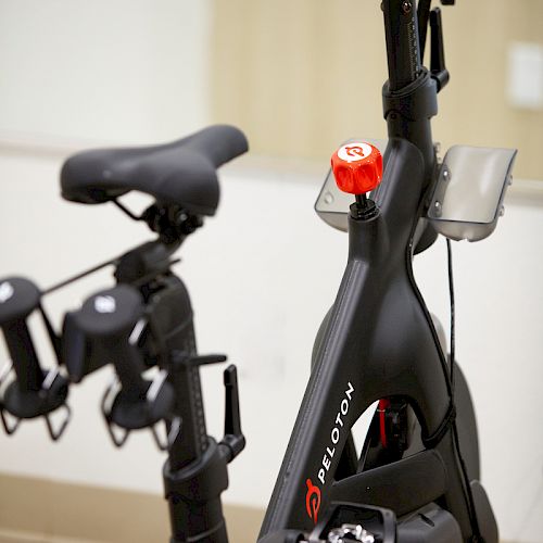 The image shows a Peloton stationary exercise bike, featuring a seat, handlebars, and a screen for virtual workout classes.