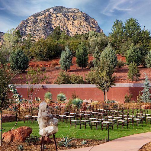 An outdoor setup with rows of chairs, surrounded by greenery and trees, with a mountain backdrop under a blue sky.