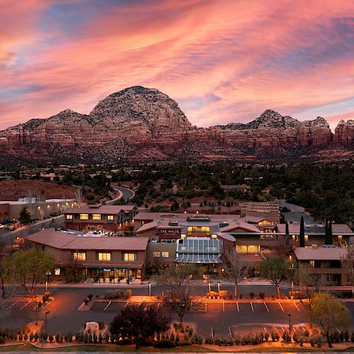 A beautiful landscape featuring a building complex in front of a mountainous backdrop under a stunning sunset sky with pink and orange clouds.