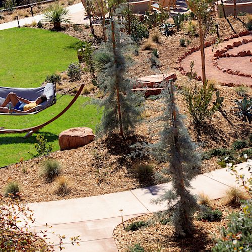 A person is relaxing on a hammock in a garden setting with pathways, plants, and a circular stone design in the background.