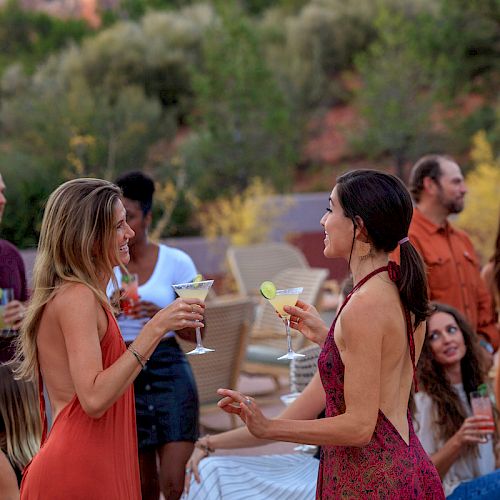 People are socializing and enjoying drinks outdoors at a gathering. Some are standing and talking, holding cocktails, while others are seated.