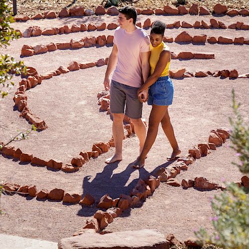 A couple walks hand in hand through a circular stone maze outdoors, surrounded by greenery, both barefoot and looking content.