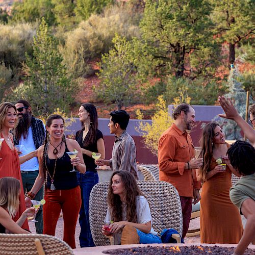 A lively outdoor gathering with people socializing, holding drinks, and enjoying themselves in a picturesque, wooded backdrop.