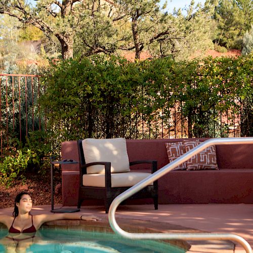 A person is relaxing in a hot tub next to a garden with trees and bushes. There is outdoor seating with cushions nearby, and the scene is peaceful.