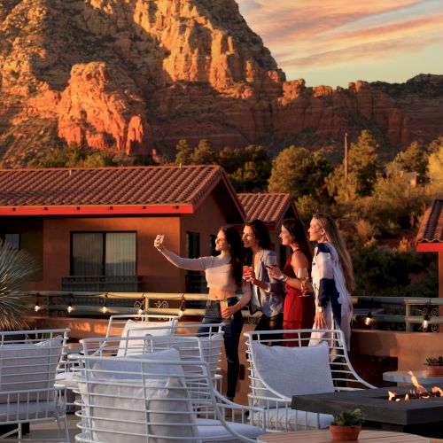 A group of people are taking a selfie on a rooftop patio with mountains in the background, during sunset.