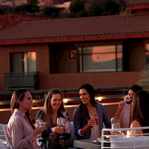 A group of five people sit outdoors at a table, drinking and smiling, with a scenic mountainous background during sunset.