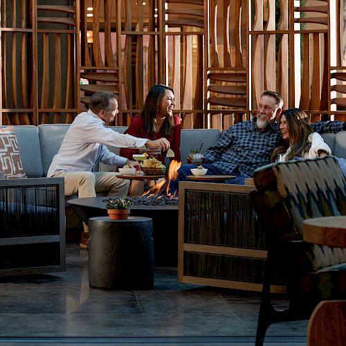 Four people are sitting in a stylish lounge area, sharing drinks and snacks around a central table, indoors. Wooden decor surrounds them.