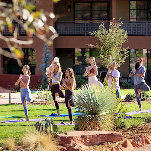 A group of people practicing yoga outdoors on mats in a grassy area with a building in the background. They are in a tree pose, balancing on one leg.