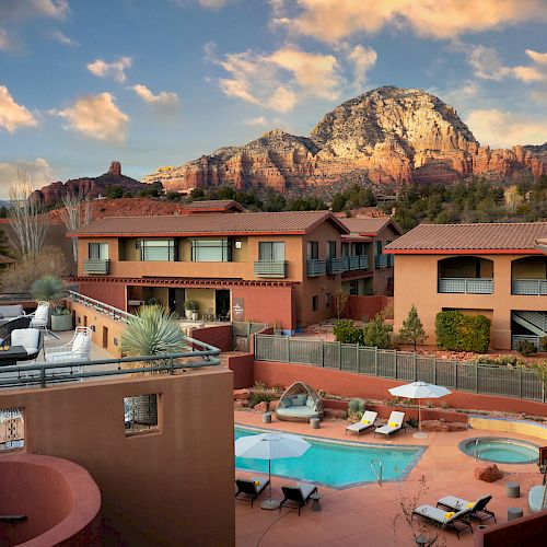 A resort with terracotta-colored buildings, a swimming pool, seating area, and mountain views under a partly cloudy sky, is depicted in the image.