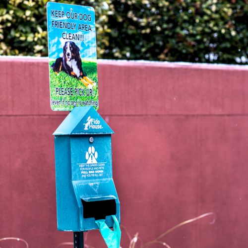 The image shows a dog waste station with a sign, dispenser for bags, and a disposal bin against a red wall and greenery in the background.