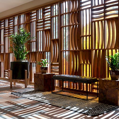 A sunlit room with wooden slats casting shadows, plants in pots, benches, and chairs arranged in a minimalist, serene setting.