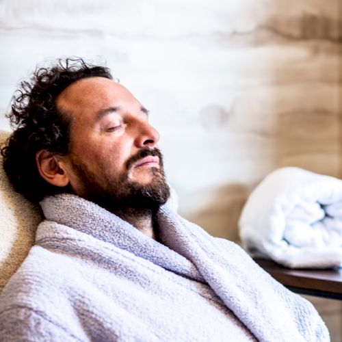 A bearded man with curly hair is relaxing in a bathrobe, eyes closed, leaning back on a cushioned chair with a rolled-up towel beside him.