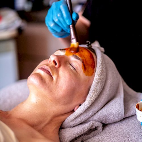 A person is receiving a facial treatment, with a cosmetologist applying a mask on their face using a brush. The person has a towel wrapped around their head.