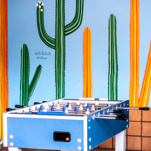 A foosball table sits in front of a colorful wall with large cacti paintings. The setting appears playful and vibrant, likely a recreational space.