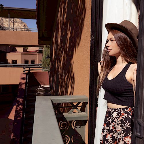 A woman in a hat stands on a balcony, gazing out at the view, with a sunny building courtyard and a distant rocky hill in the background.