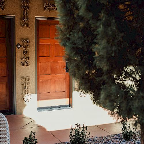 The image shows two wooden doors with intricate decorations on the wall beside them. In the foreground, there's a tree and a small garden area with plants.