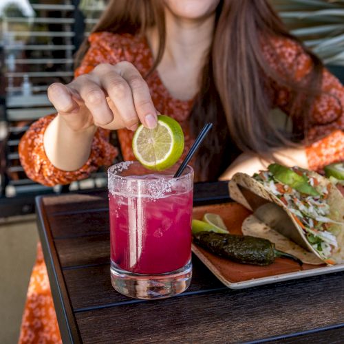 A person is squeezing a lime into a pink drink with a straw, and a plate of tacos is on the table. The person is wearing an orange patterned top.